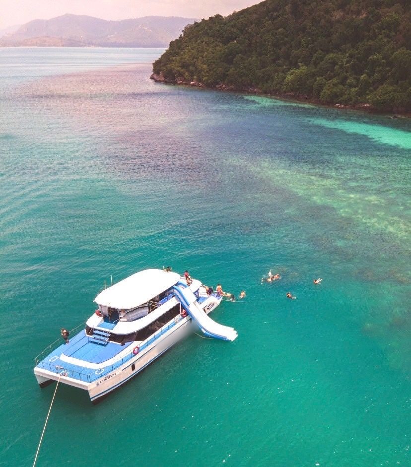 Catamaran with a water slide anchored in clear water, guests swimming beside it